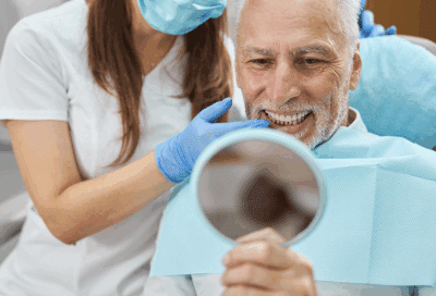 older man looking at his brand new teeth in mirror while dentist looks with him, representing All-on-X dental implants