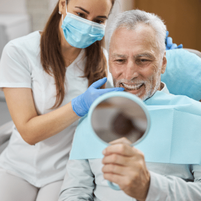 Older man looking at his brand new teeth in mirror while dentist looks with him, representing all-on-x dental implants