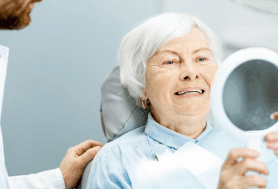 Older woman looking in a small mirror at her new teeth, representing dental implants in Lansdowne VA
