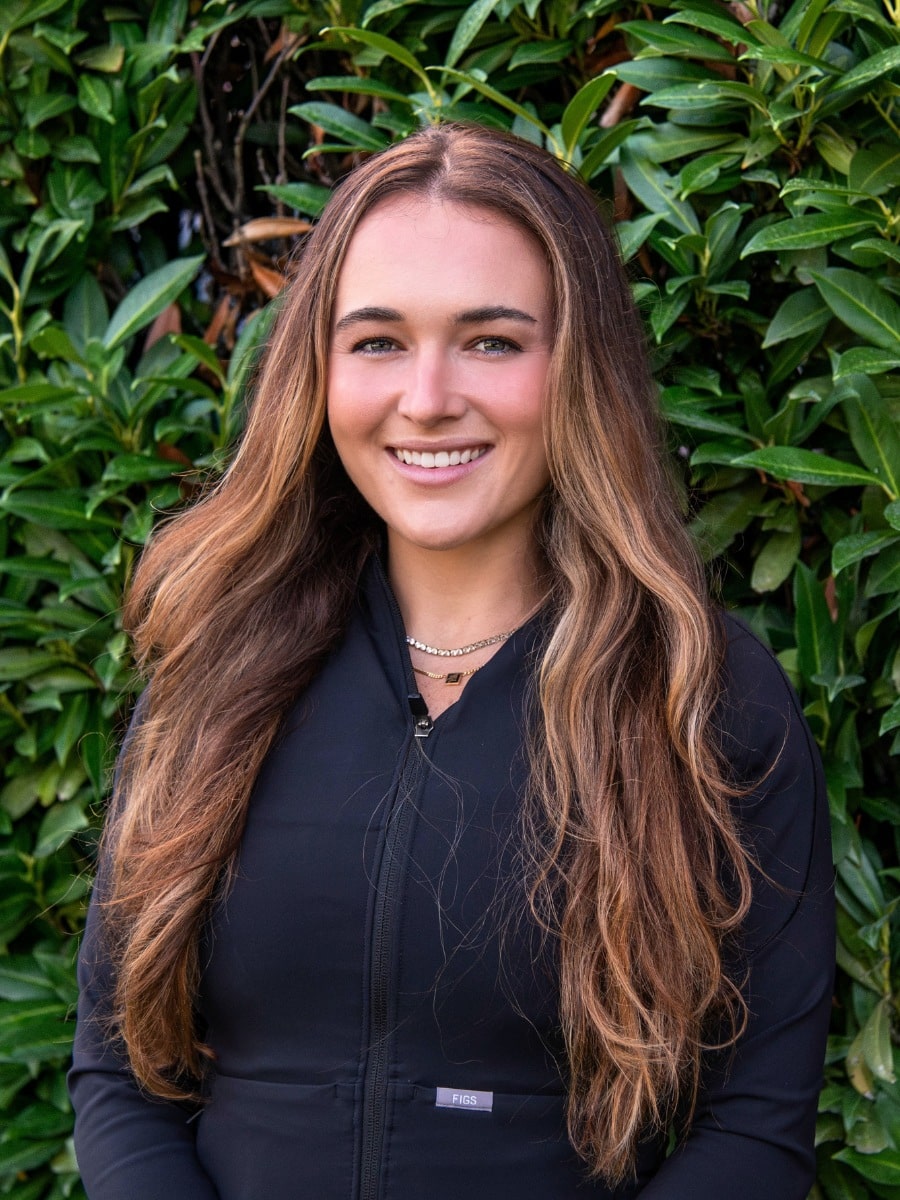 Smiling woman with long wavy hair wearing black scrubs, standing outdoors in front of green foliage.