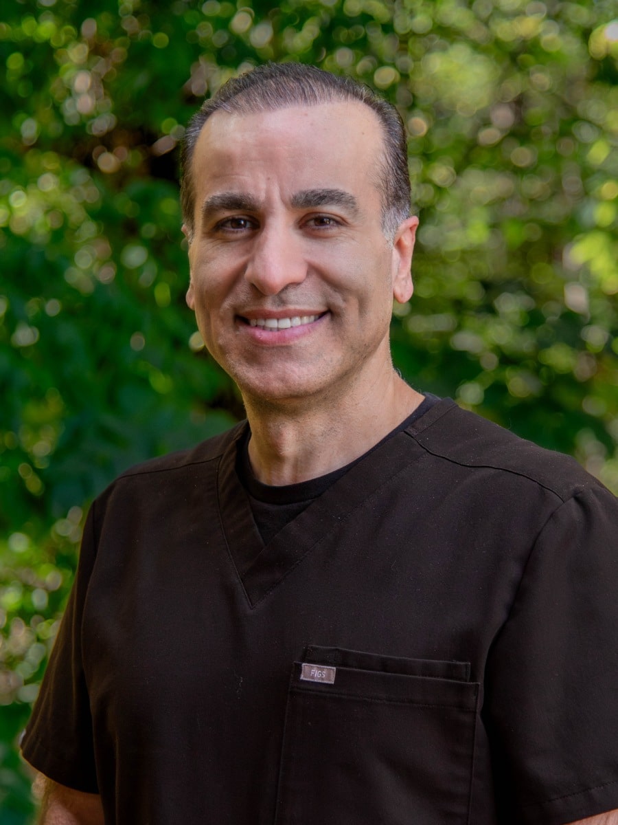 Man wearing black scrubs smiling outdoors in front of green foliage.