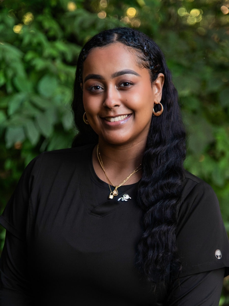 Smiling woman in black scrubs standing outdoors in front of green foliage.