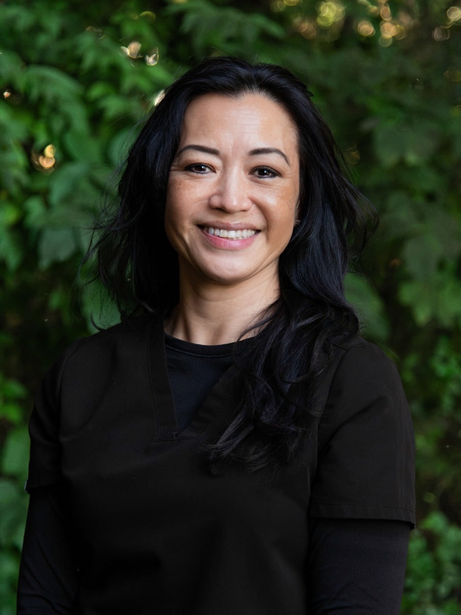 Smiling woman in black scrubs standing outdoors in front of green foliage.